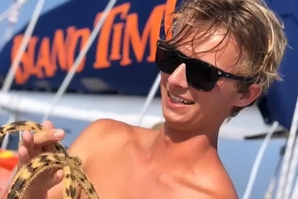 A smiling young man wearing sunglasses is holding a starfish in front of a boat named Island Time