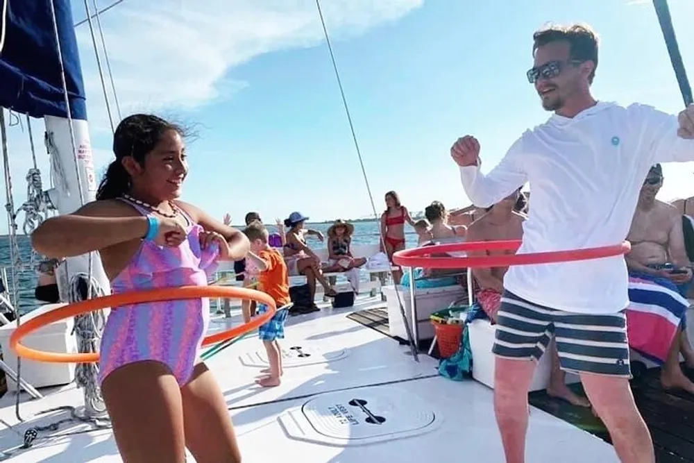 A girl and a man are enjoying hula hooping on the deck of a sailboat with other passengers in the background under a clear blue sky