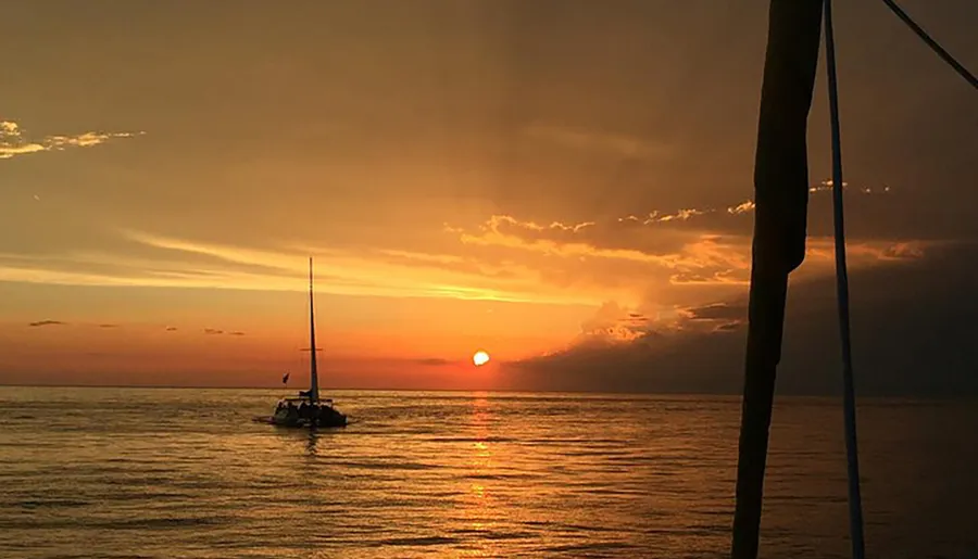 A sailboat is silhouetted against a stunning sunset over the ocean, with vibrant oranges and yellows coloring the sky and reflecting on the water.