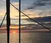 A sailboat with a black sail bearing a pelican image is cruising on the water at sunset with a group of passengers enjoying the scenery