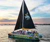 A sailboat with a black sail bearing a pelican image is cruising on the water at sunset with a group of passengers enjoying the scenery