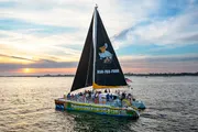 The image shows a colorful catamaran with a group of people on board, sailing on calm waters during a beautiful sunset.