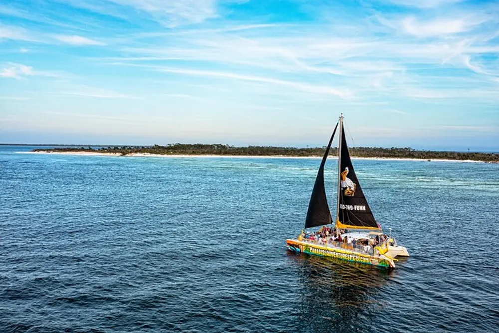 A catamaran with a brightly colored hull and a black sail filled with people is sailing in blue waters near a coastline