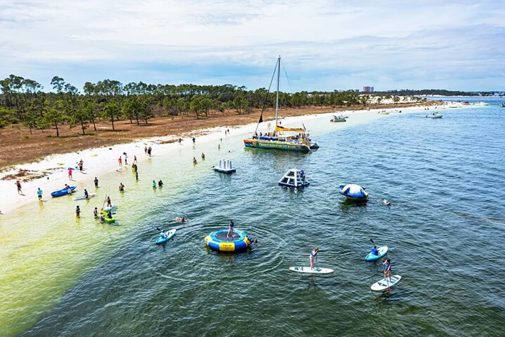 A busy beach scene with people engaging in various water activities such as stand-up paddleboarding swimming and relaxing on floating rafts with a sailboat anchored nearby and a backdrop of trees and a clear sky