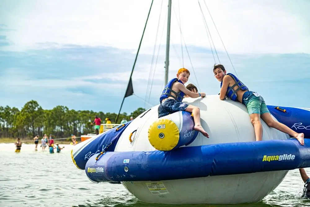 Two children wearing life jackets are sitting on a large inflatable water toy with the backdrop of a lake and partially visible sailboat mast