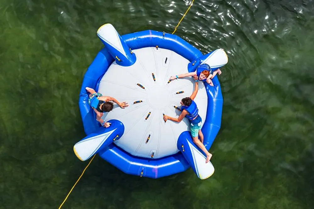The image shows an aerial view of three people lounging on a large round inflatable water raft with blue and white colors floating on a body of water
