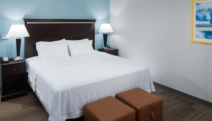 This image shows a neatly made bed in a modern hotel room with two bedside tables lamps and a framed picture on the wall conveying a clean and simple aesthetic