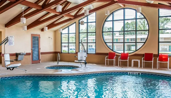 The image shows an indoor swimming pool with a hot tub surrounded by chairs and overlooked by a large circular window inside a room with a vaulted ceiling and exposed wooden beams
