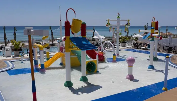 A colorful childrens water playground with slides and fountains is set against a backdrop of a sandy beach and the ocean