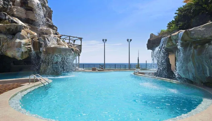 An outdoor pool with a cascading artificial waterfall overlooking the sea set against a clear sky