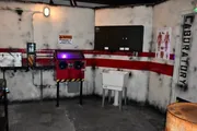 The image depicts an industrial-themed room designed to resemble a laboratory or hazardous area, complete with warning signs, a safety tape barrier, a sink, and a red machine or console.