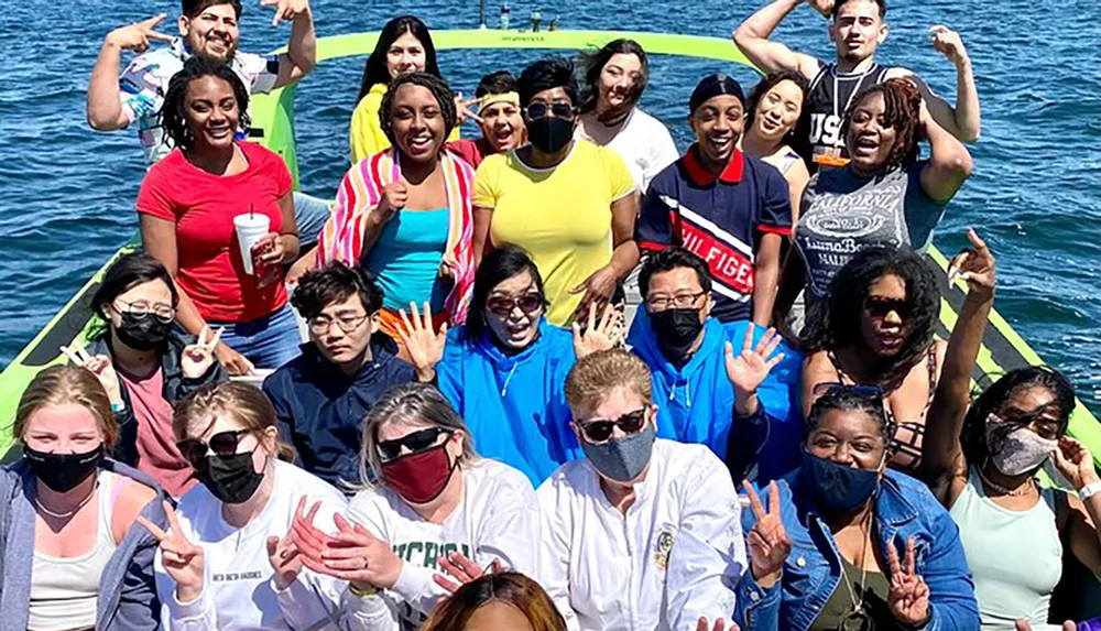 A diverse group of smiling people are waving and posing for a photo on a boat with some wearing face masks