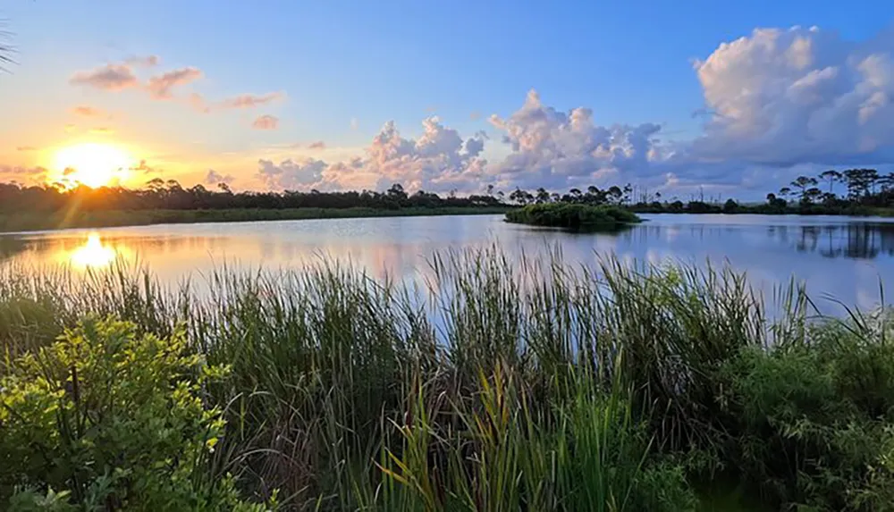 The image shows a serene sunset over a calm lake surrounded by lush greenery with clouds in the sky reflecting on the waters surface