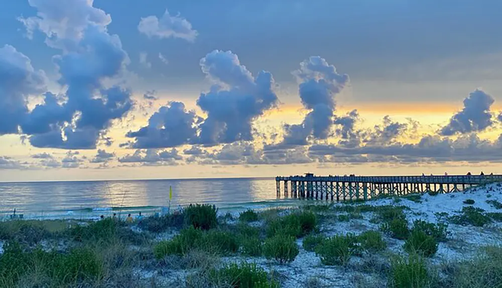 The image captures a tranquil sunset over a beach with a wooden pier extending into the calm sea under a sky filled with dramatic clouds