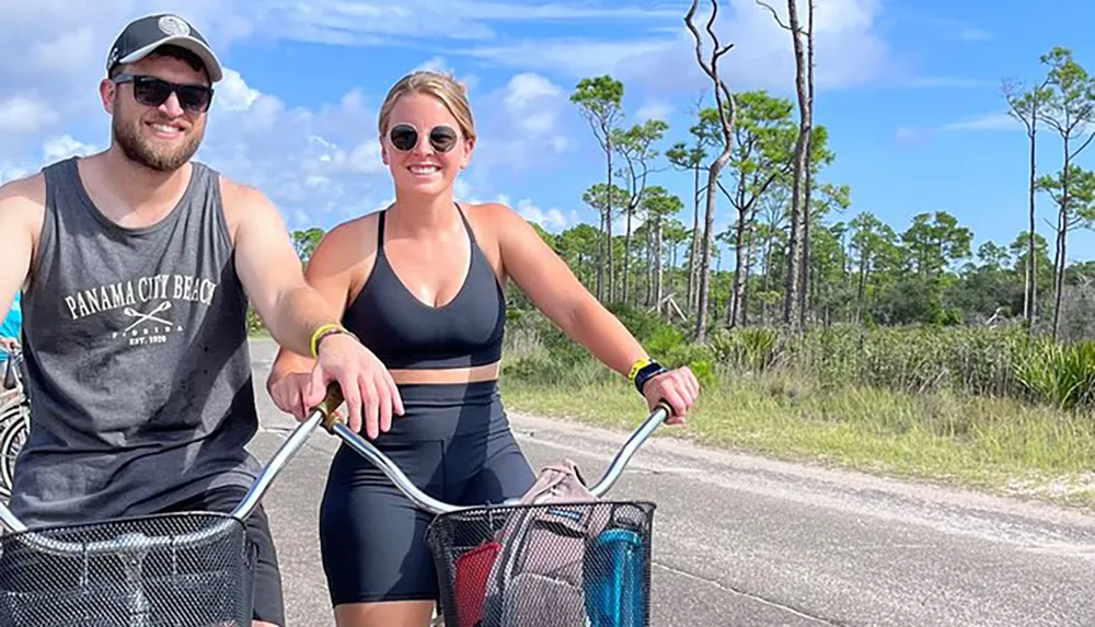 A smiling man and woman are riding bicycles on a sunny day along a path flanked by trees and grass