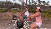 Two people are smiling while riding bicycles on a path through a natural landscape during twilight.