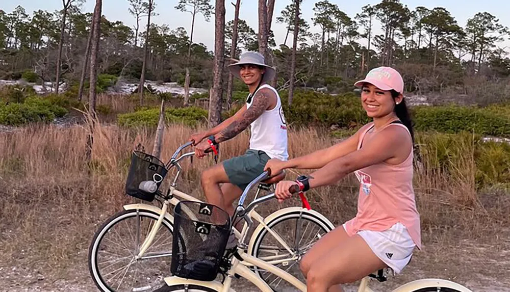 Two people are smiling while riding bicycles on a path through a natural landscape during twilight