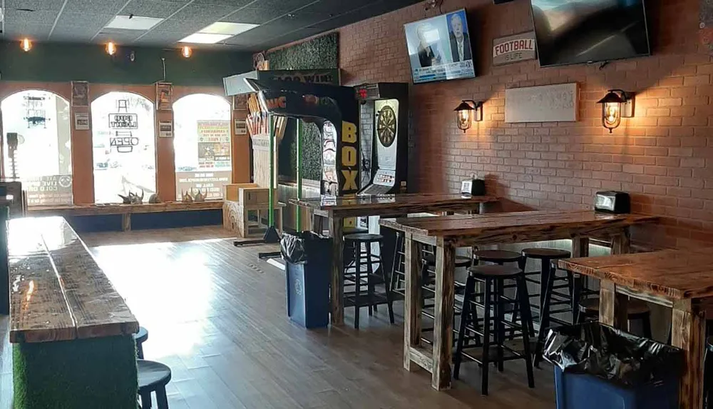 The image shows the interior of a casual bar with empty wooden tables and chairs a jukebox a dartboard and televisions displaying a sports channel creating a relaxed atmosphere for socializing and entertainment