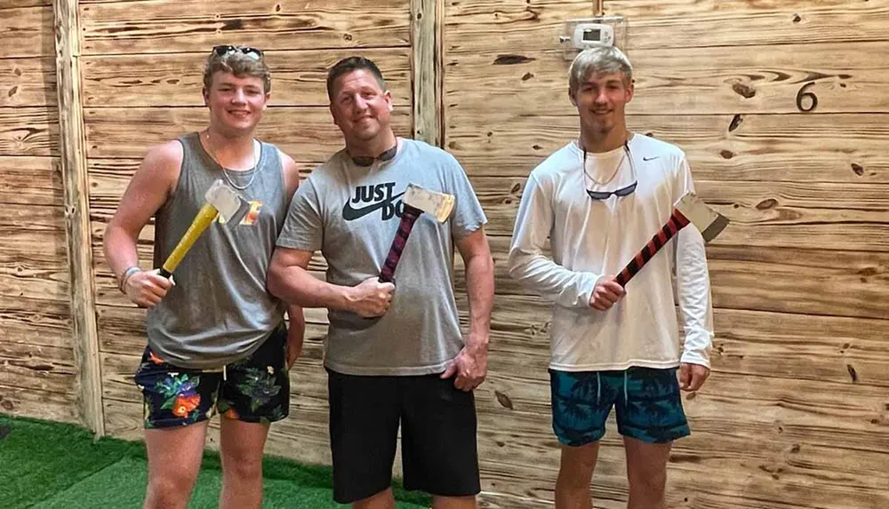 Three individuals are posing with axes in front of a wooden wall likely at an axe-throwing venue