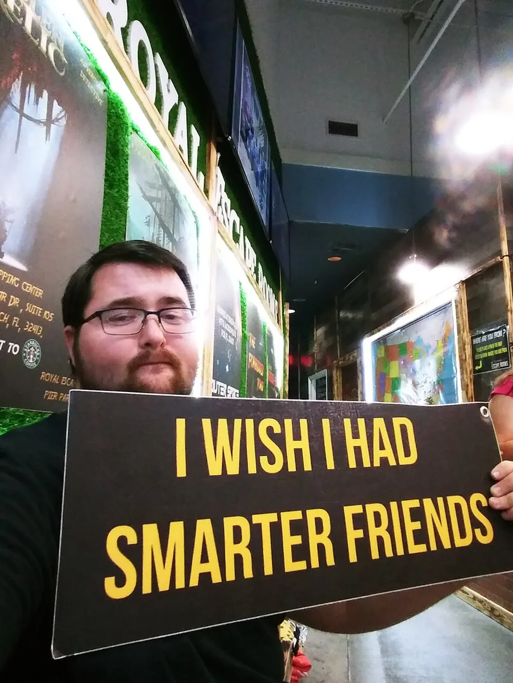 A person is taking a selfie while holding a sign that reads I WISH I HAD SMARTER FRIENDS