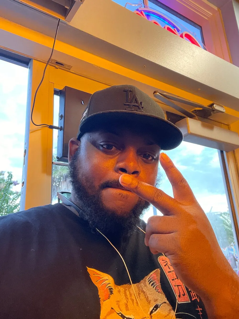 A person wearing a baseball cap is taking a selfie while making a peace sign with fingers on one hand and touching their nose with the other