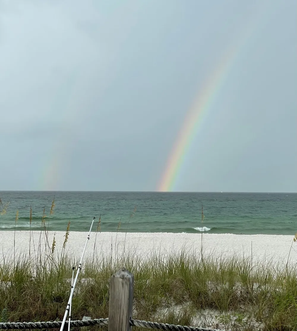 A vibrant rainbow emerges from a cloudy sky above a tranquil white sand beach