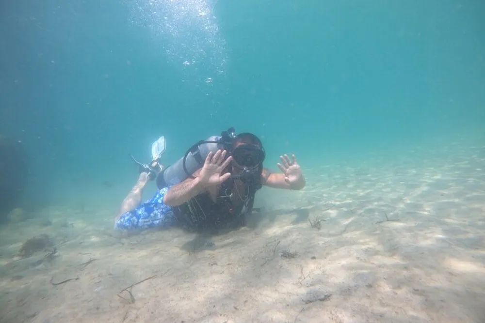 A scuba diver is kneeling on the sandy ocean floor while underwater making a playful gesture with hands spread towards the camera