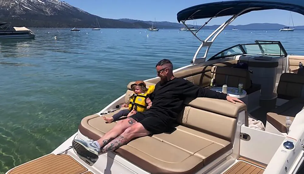 An adult and a child are enjoying a sunny day on a boat with a view of a calm lake and mountains in the background