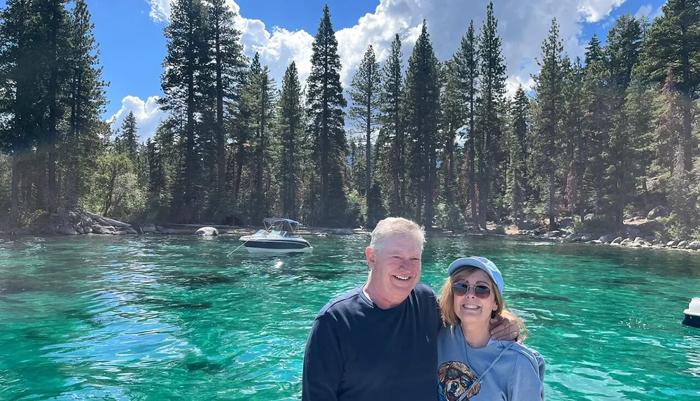 A smiling couple poses for a photo with a backdrop of crystal-clear turquoise water and a lush pine forest under a blue sky