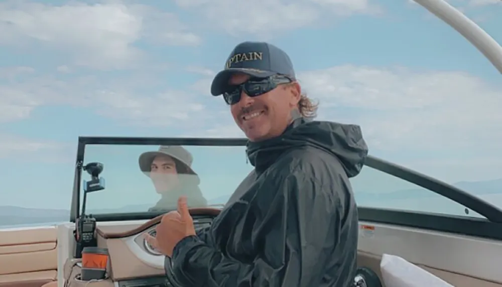 A smiling person wearing a cap labeled CAPTAIN is giving a thumbs-up while steering a boat with another persons reflection visible in the windscreen