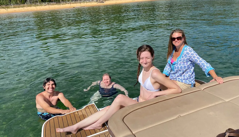 Four individuals are enjoying a sunny day on a boat and in the water with crystal clear waters and a sandy shore in the background