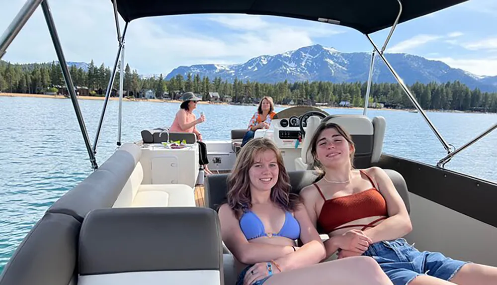 Two people are smiling at the camera while relaxing on a boat with a mountainous backdrop and two others are sitting towards the rear enjoying the lake view