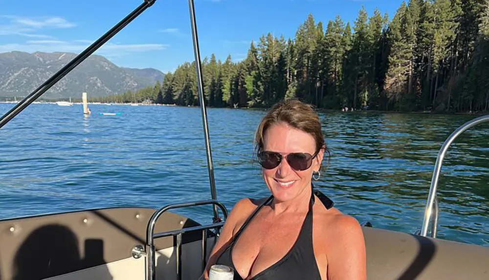 A woman in sunglasses is smiling on a boat with a backdrop of clear water and forested mountains