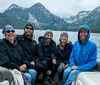 A group of five people wearing warm clothes and smiles are seated on a boat with a scenic backdrop of a mountainous forested area and overcast skies