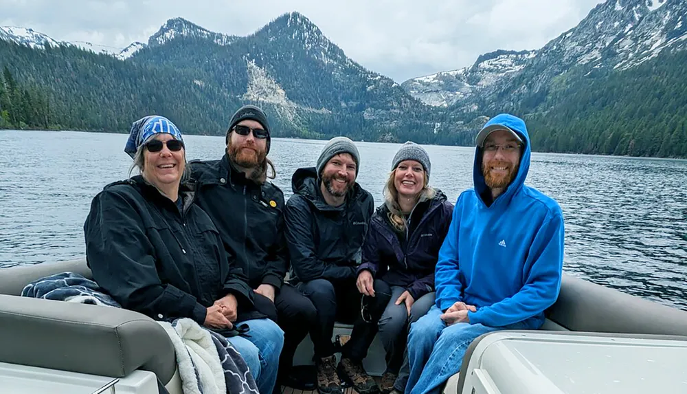 A group of five people wearing warm clothes and smiles are seated on a boat with a scenic backdrop of a mountainous forested area and overcast skies