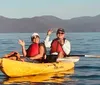 A group of people wearing life vests are smiling and paddling in tandem yellow kayaks on a lake with a backdrop of forested hills and clouds