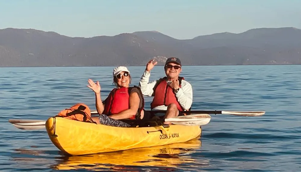 Two people are smiling and waving from a yellow tandem kayak on calm blue waters with mountains in the background under a clear sky