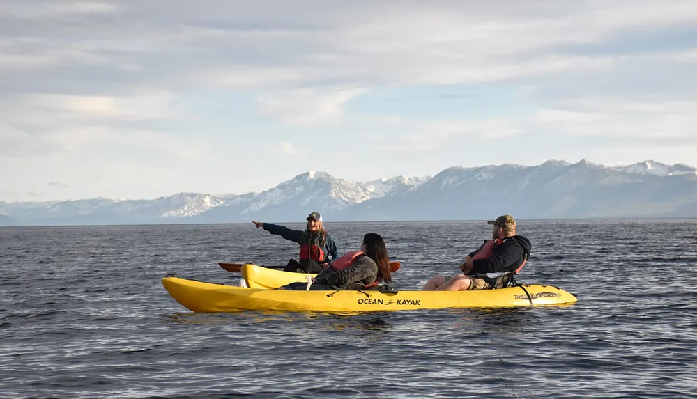 Three people are kayaking on a calm sea with one person pointing towards the horizon against a backdrop of distant mountains
