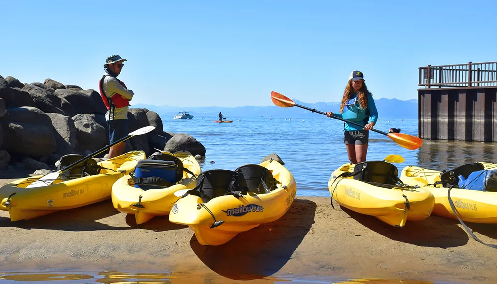 Two individuals in outdoor gear stand by yellow kayaks on a sandy shore with a clear blue lake and mountainous background