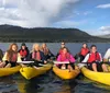 A group of people wearing life vests are smiling and paddling in tandem yellow kayaks on a lake with a backdrop of forested hills and clouds