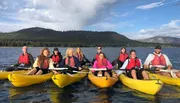 A group of people wearing life vests are smiling and paddling in tandem yellow kayaks on a lake with a backdrop of forested hills and clouds.