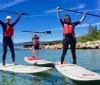 Three people in wetsuits are striking triumphant poses while standing on paddleboards in a clear blue lake with a scenic mountainous backdrop