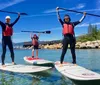 Three people in wetsuits are striking triumphant poses while standing on paddleboards in a clear blue lake with a scenic mountainous backdrop