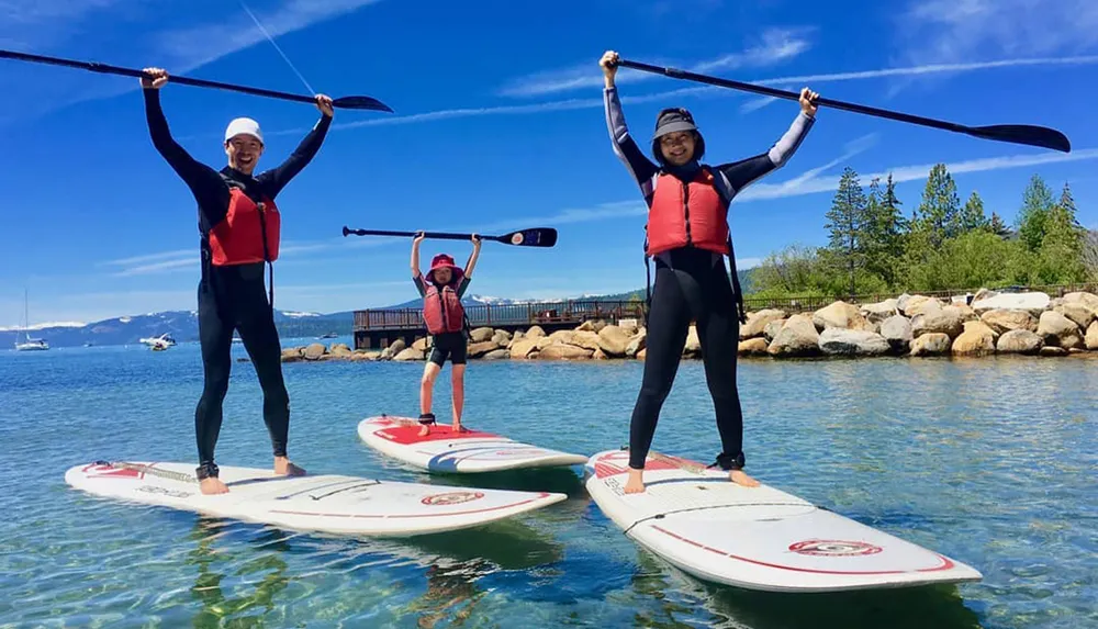 Three people in wetsuits are striking triumphant poses while standing on paddleboards in a clear blue lake with a scenic mountainous backdrop