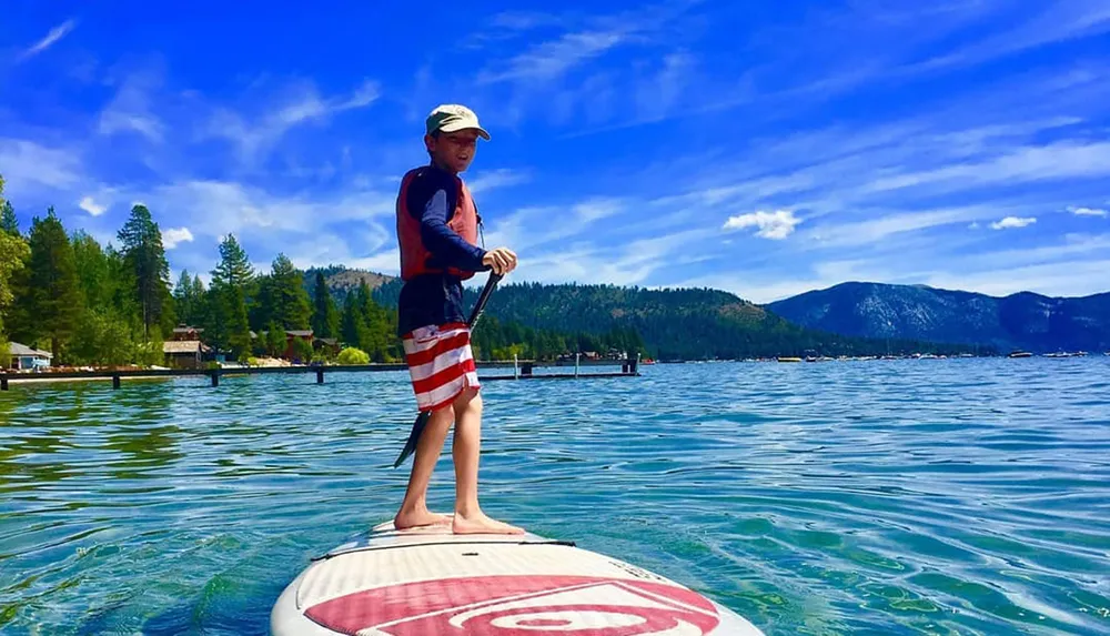 A child is standing on a paddleboard on a clear blue lake with mountains in the background