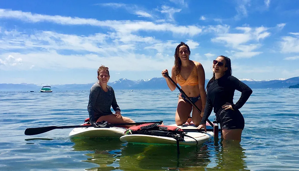 Three people are standing and sitting on paddleboards on a calm blue lake with mountains in the distance under a partly cloudy sky