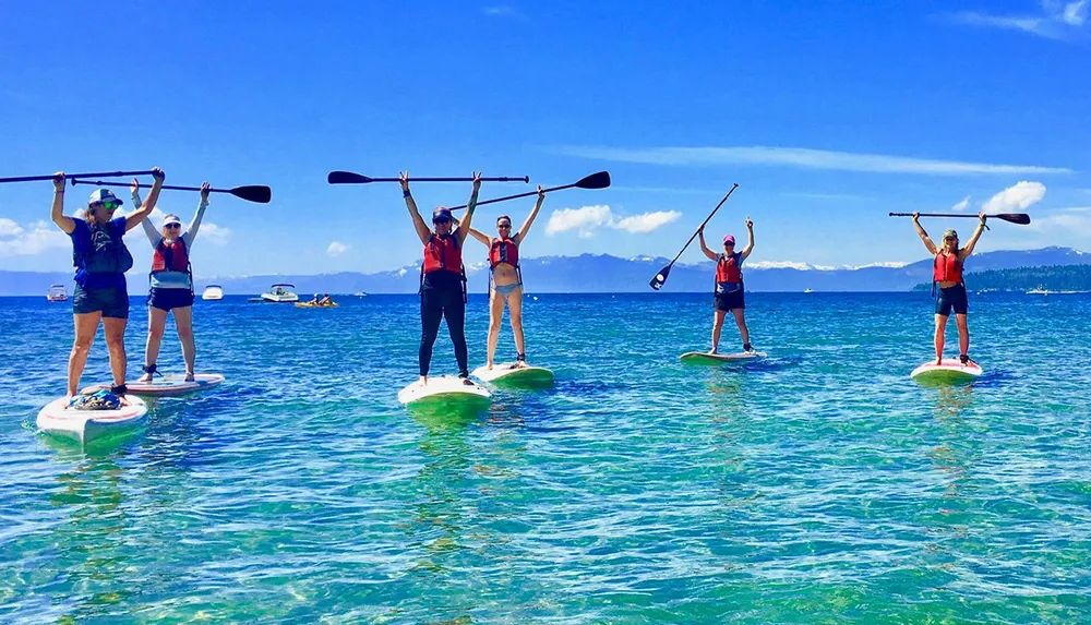 A group of people is standing on paddleboards on a clear blue water body holding their paddles up in the air under a sunny sky