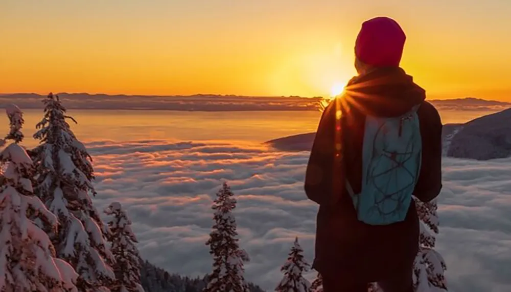 A person in winter clothing stands on a snow-covered mountain gazing at a beautiful sunrise above a sea of clouds