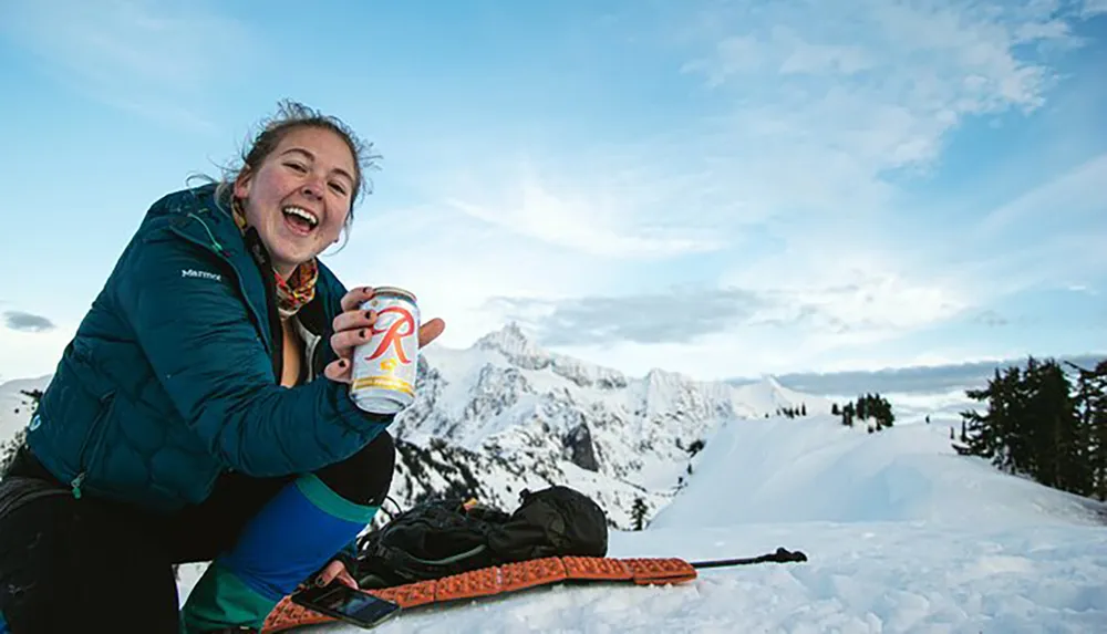 A cheerful person is holding a drink can while seated in the snow with skis nearby against a backdrop of mountainous terrain