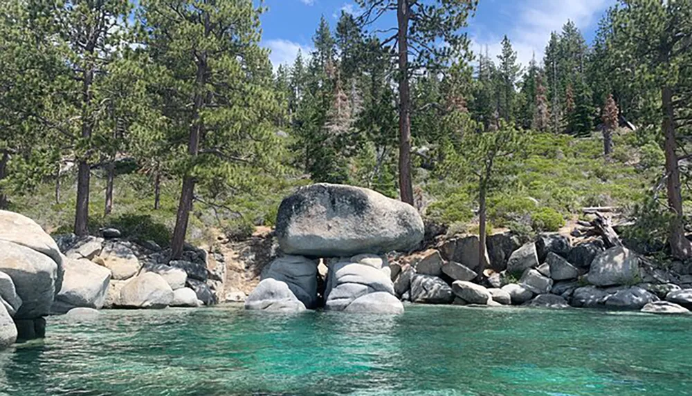The image shows a large boulder precariously balanced on top of several other rocks near the edge of a clear blue water body against a backdrop of a coniferous forest